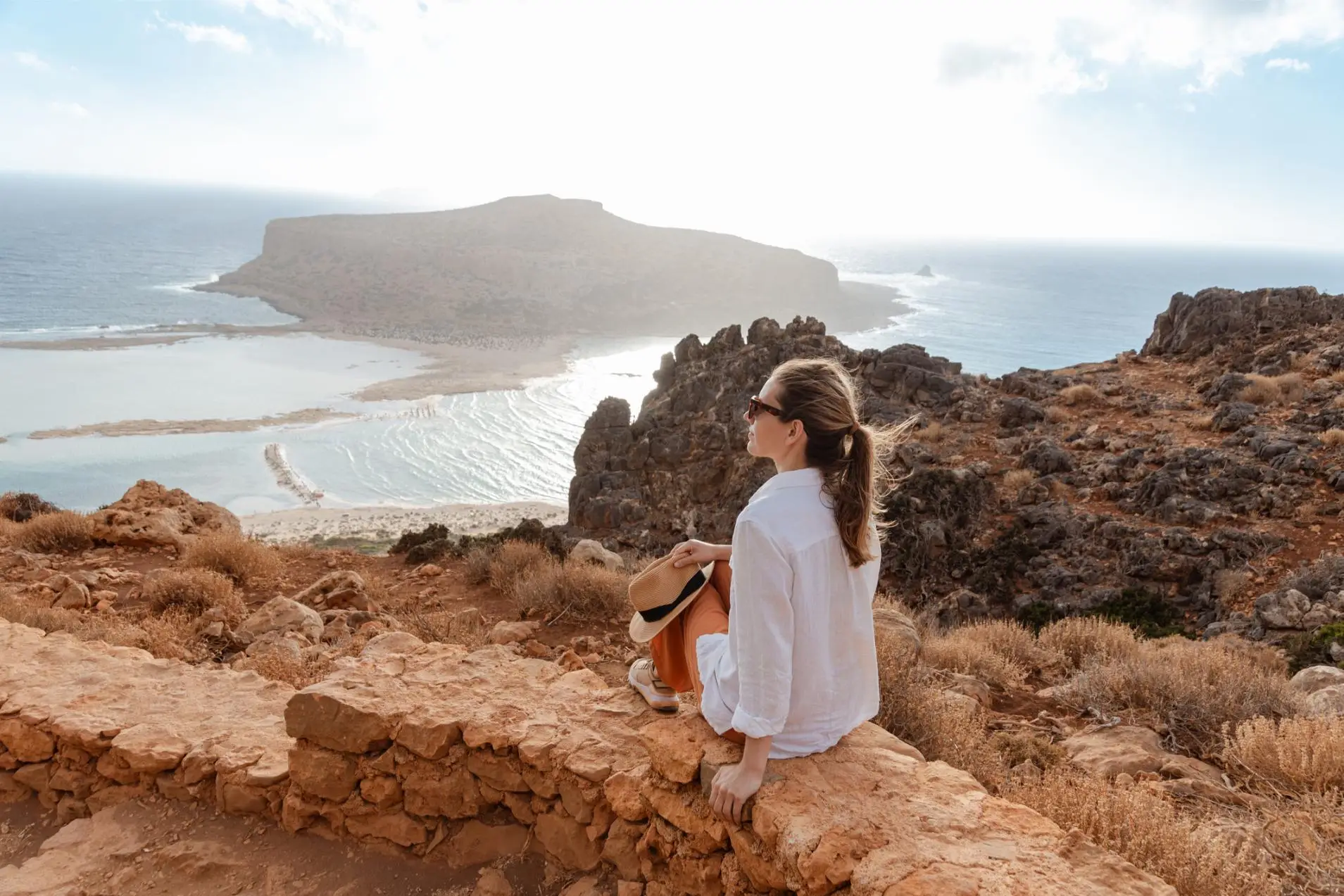 A woman relaxes and admires the scenic coastal view from a rocky lookout on a bright sunny day.